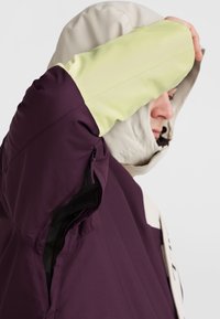 Wasserabweisende Jacke in Lila mit Creme- und Limettengrün-Akzenten. Verfügt über eine Kapuze, eine Reißverschlusstasche und einen strukturierten Stoff.