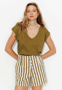 T-shirt vert olive à col en V avec des manches courtes, associé à un short taille haute avec des rayures verticales blanches et marron et des détails de boutons.