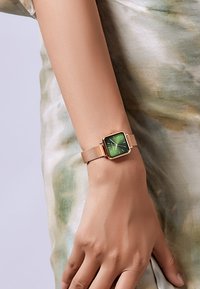 Zeleni sat s kvadratnim brojčanikom i zlatnom mrežicom, sa minimalističkim oznakama sati i elegantnim dizajnom na zapešću protiv prigušene tkanine.