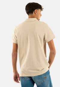 Krémová bavlněná polokošile s krátkým rukávem, detailem větrání na zadní straně a volným střihem, doplněná modrými džínami.