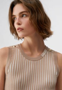 Femme aux cheveux courts bruns portant un haut sans manches beige et blanc à rayures verticales avec des boutons dorés sur les épaules, regardant vers sa gauche.