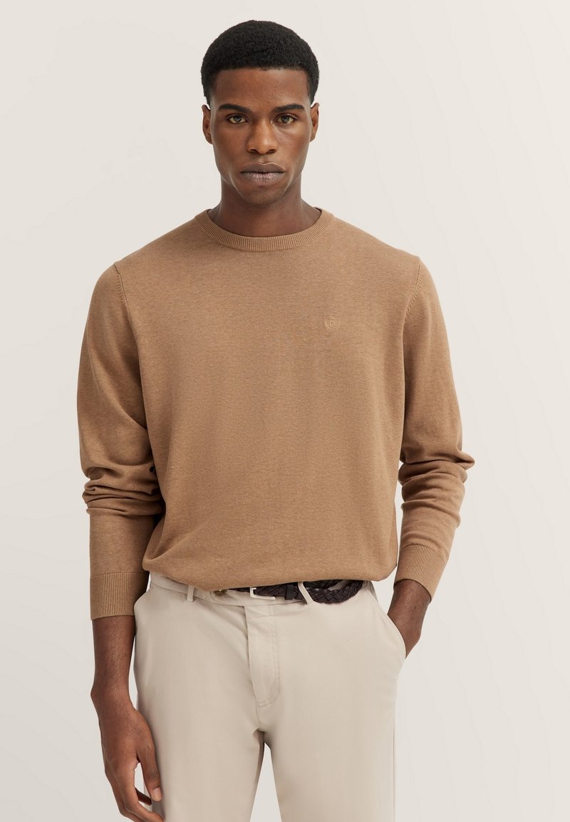 Brauner Strickpullover mit Rundhalsausschnitt und langen Ärmeln, ausgestattet mit gerippten Bündchen und Saum, kombiniert mit hellbeigen Hosen. Subtiles Logo-Detail.