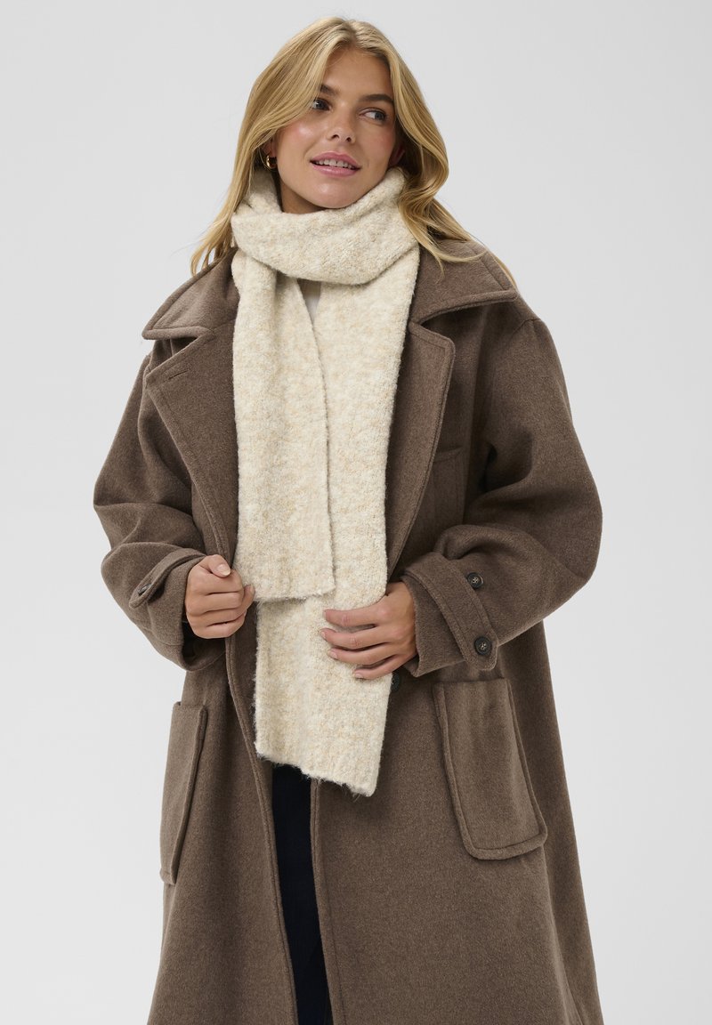 Kvinna i brun ullkappa och en krämfärgad stickad scarf, som tittar åt sidan med ett lätt leende, händerna justerar kappan.