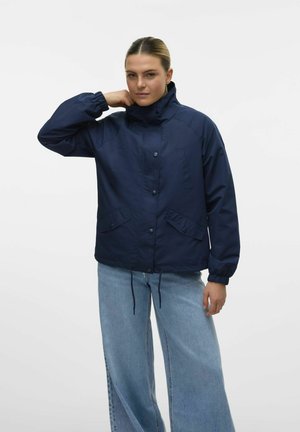 Veste coupe-vent bleu marine avec un col montant, des boutons-pression, des poignets élastiques et des poches latérales ; portée avec un jean large bleu clair.