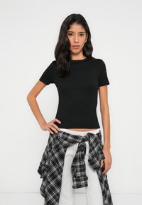 Μαύρο κοντομάνικο εφαρμοστό μπλουζάκι από ριμπ ύφασμα. Συνδυάζεται με άνετο γκρι παντελόνι και καρό πουκάμισο σε μαύρο και άσπρο, δεμένο γύρω από τη μέση.