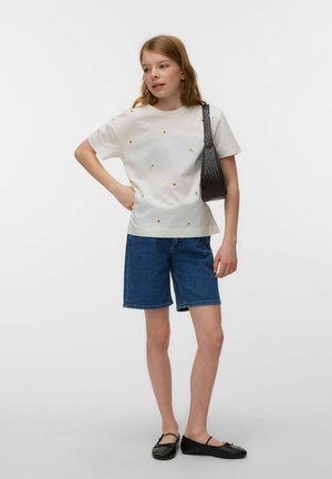 Young woman wearing white floral t-shirt, knee-length denim shorts, black flats, and carrying a black shoulder bag, standing with hand on hip.