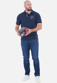 Homme barbu portant un polo marine et un jean tient un ballon de rugby, regardant légèrement sur sa gauche contre un fond blanc uni.