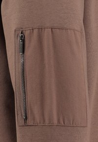 Tessuto marrone con una trama liscia che presenta una tasca laterale con dettaglio zipper. La tasca è cucita sul capo, enfatizzando il suo design.