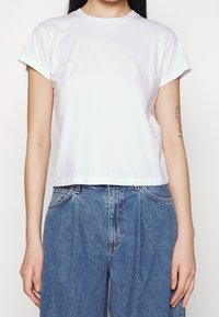 Vit bomull t-shirt med avslappnad passform, korta ärmar och en något beskuren nederkant. Den har en subtil präglad logga över bröstet.