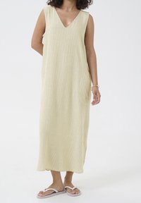 Robe sans manches jusqu'aux genoux, de couleur jaune clair avec de fines rayures blanches, dotée d'un décolleté en V et de poches latérales, portée avec des tongs blanches.