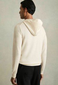 Beige Kapuzenpullover mit langen Ärmeln, strukturiertem Stoff, gerippten Bündchen und lässiger Passform, kombiniert mit dunklen Hosen.