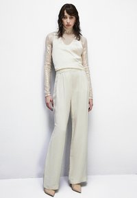 Top in satin avorio con scollo a V, abbinato a pantaloni a gamba larga a vita alta. Maniche lunghe in pizzo trasparente e con paillettes aggiungono texture. Tacchi a punta neutri.