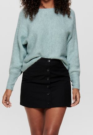 Femme portant un pull côtelé bleu clair rentré dans une mini-jupe noire taille haute à boutons sur le devant, sur un fond uni.