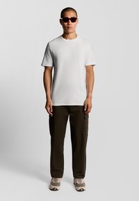 Man in een donker zonnebril, witte korte mouwen T-shirt, donkere cargobroek en beige sneakers staat tegen een effen grijze achtergrond.