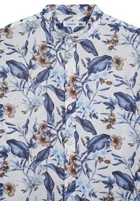 Camicia a maniche corte con bottoni in tessuto leggero con sfondo bianco. Presenta motivi floreali blu e marroni, colletto a sbuffo e bottoni bianchi.