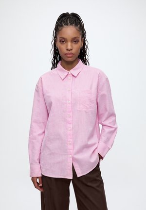 Femme aux cheveux tressés portant une chemise rayée rose et blanche boutonnée et un pantalon marron, debout devant un fond clair uni.