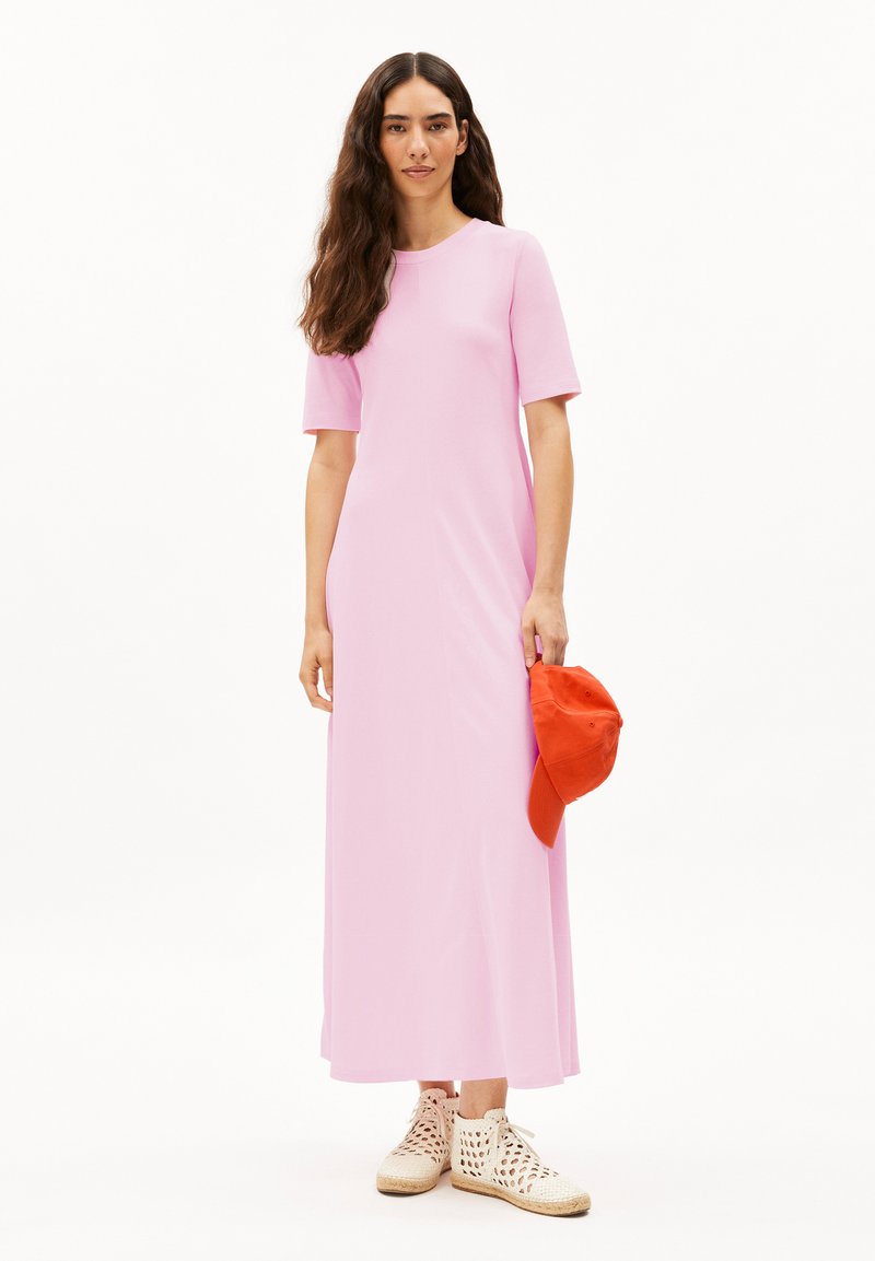Pink, short-sleeve maxi dress with a round neckline. Model holds an orange cap, wearing beige, perforated footwear. Simple design and smooth texture.