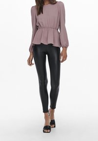Blouse mauve à volants avec taille élastique, manches longues ; associée à des leggings noirs en simili cuir serrés et des talons noirs ouverts à l'avant.