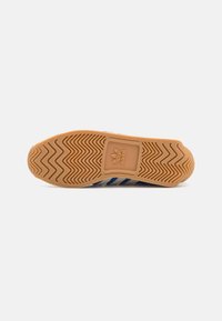 Gumová podrážka boty Adidas se zigzagovým vzorem a logem Adidas uprostřed, viděná zespodu na bílém pozadí.