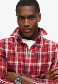 Camicia a quadri rossa e blu navy con colletto abbottonato, caratterizzata da una tasca frontale e un piccolo marchio del brand. Il tessuto appare testurizzato.