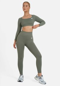 Maslinasto zeleni sportski crop top i tajice visokog struka izrađene od tekstuiranog materijala s bijelim logotipom, uparene s svijetlosivim sportskim cipelama.