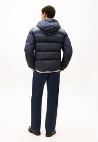Marineblauer Daunenjacke mit Kapuze, gestepptem Design und elastischen Bündchen, kombiniert mit marineblauen Hosen und schwarzen Schuhen, von hinten betrachtet.