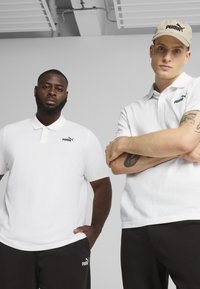 Due modelli indossano polo bianche con il logo Puma nero, abbinate a pantaloni neri. L'ambientazione presenta uno sfondo minimalista.