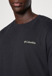 Férfi fekete kereknyakú pólót visel, amelyen bal mellkason egy kis világos színű Columbia logó található, egy sima háttér előtt.
