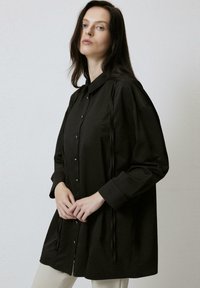 Black long-sleeve shirt with a button-down front, featuring a loose fit, subtle pleats, and ribbon accents along the sides.