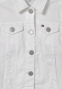 Valkoinen farkkutakki, jossa on kaksi rintataskua, hopeiset napit ja kontrastivärinen ompelu. Vasemmassa taskussa on pieni monivärinen logo.