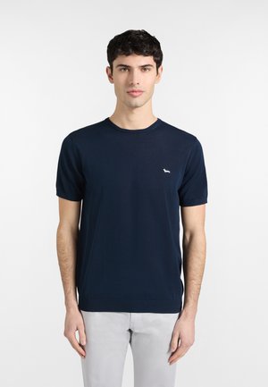 Mladý muž s krátkymi tmavými vlasmi, oblečený v tmavomodrej košeli s krátkym rukávom s malým bielym logom psa a svetlosivými nohavicami, smerujúci vpred.