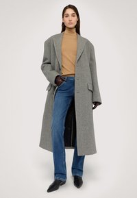 Abrigo oversized de mezcla de lana gris, suéter de cuello alto beige, jeans azules y botas de tobillo negras con punta afilada. Textura suave con acentos mínimos de herrajes.