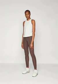 Vékony, ujjatlan fehér felső barna leggings-szel és fehér sportcipőkkel párosítva. Sima, ruganyos anyag és testhezálló dizájn.