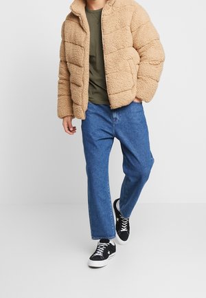 Beige, flauschige Pufferjacke mit Reißverschluss, strukturiertem Stoff und horizontaler Steppung, kombiniert mit blauen geraden Jeans und schwarzen Sneakers.