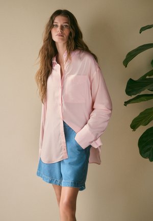 Kvinde med langt bølget brunt hår iført en løs, lys rosa skjorte med knapper og blå denimshorts, der står ved en grøn plante mod en beige væg.