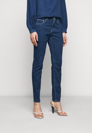 Kobieta ubrana w ciemnoniebieskie dżinsy o dopasowanym kroju, granatową bluzkę z długim rękawem oraz białe sandały na wysokim obcasie, stojąca na jednolitym tle.