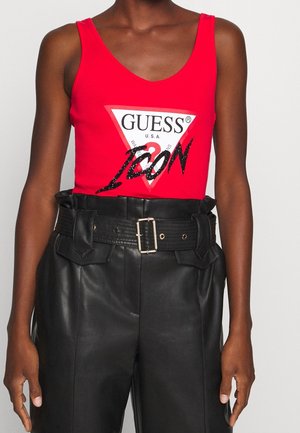 Persoon draagt een rood mouwloos topje met het "GUESS ICON"-logo en een zwarte leren broek met hoge taille, voorzien van een brede riem en metalen gesp.