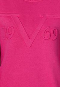 Różowa bluza z podniesionymi literami "V" i "1969" na przodzie, o gładkiej strukturze i okrągłym dekolcie.