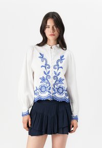 Blusa bianca con ricamo floreale blu, dotata di colletto arricciato e chiusura con bottoni. Maniche lunghe e orlo a scallop. Abbinata a una gonna blu navy.