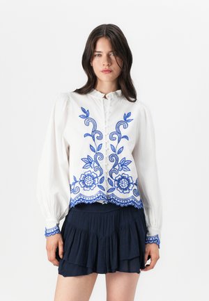 White blouse with blue floral embroidery, featuring a ruffled collar and button closure. Long sleeves and a scalloped hem. Paired with a navy skirt.