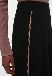 Zbliżenie osoby w czarnej, prążkowanej spódnicy z pionowym beżowym przeszyciem oraz długim rękawem teksturowanej bluzki w kolorze mauve.