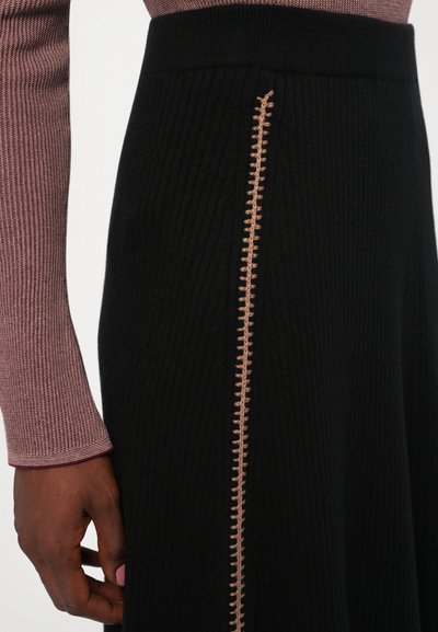 Close-up of a person wearing a black ribbed skirt with vertical beige stitch detail and a long-sleeve textured mauve top.
