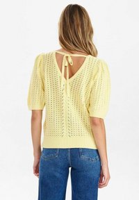Stickad gul tröja med korta puffärmar, V-ringad bak och en knytdetalj. Har mönster med öppen vävning och ribbad fåll.