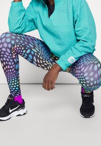 Pullover turchese texturizzato sopra una maglietta nera, abbinato a leggings a pois multicolori e scarpe da corsa nere con dettagli bianchi.