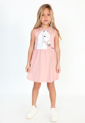 Ragazza giovane con lunghi capelli biondi che indossa un vestito rosa senza maniche con una grafica di un cavallo bianco, abbinato a scarpe da ginnastica bianche.
