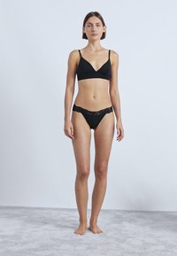 Schwarzes Dessous-Set mit einem Bralette mit dünnen Trägern und Spitzenbesatz an den Slips, dargestellt an einem Modell vor einem hellen Hintergrund.