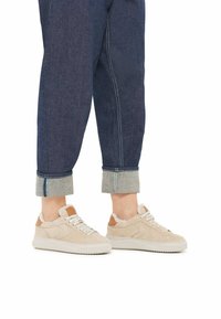 Beige Wildleder-Sneaker mit weißer Gummisohle, Schnürdesign und braunen Lederelementen. Geführt werden sie mit dunkelblauen, hochgeschlagenen Jeanshosen mit grauer Einfassung.