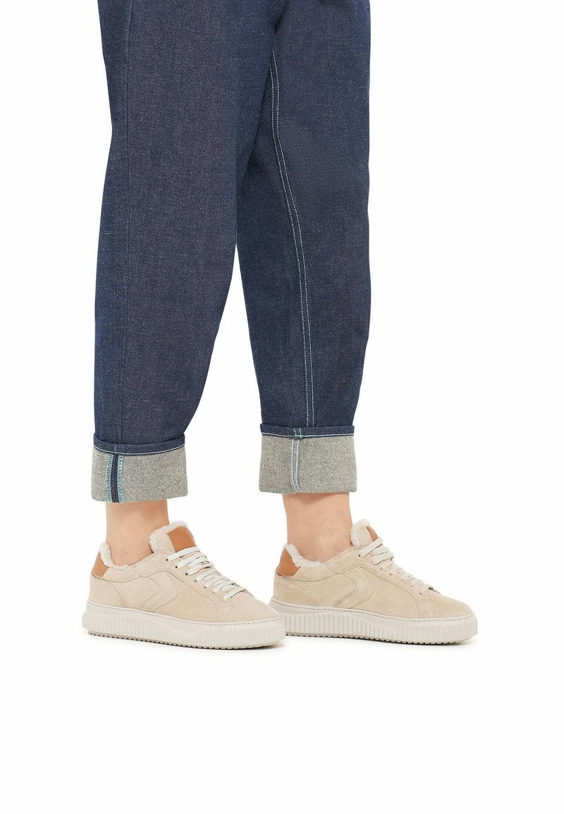 Beige Wildleder-Sneaker mit weißer Gummisohle, Schnürdesign und braunen Lederelementen. Geführt werden sie mit dunkelblauen, hochgeschlagenen Jeanshosen mit grauer Einfassung.