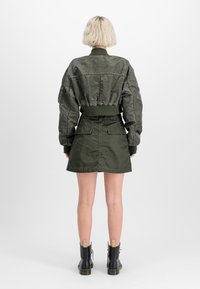 Olijfgroene bomberjack met ribgebreide boorden, gecombineerd met een bijpassende groene A-lijn rok en zwarte veterboots met enkelhoge schacht. Textuurdetails van de stof zijn zichtbaar.