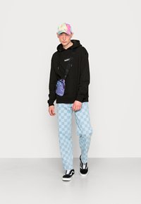 Felpa con cappuccio nera, pantaloni a scacchi blu chiaro, sneakers bianco e nero, cappellino tie-dye pastello, e una piccola borsa a tracolla viola.
